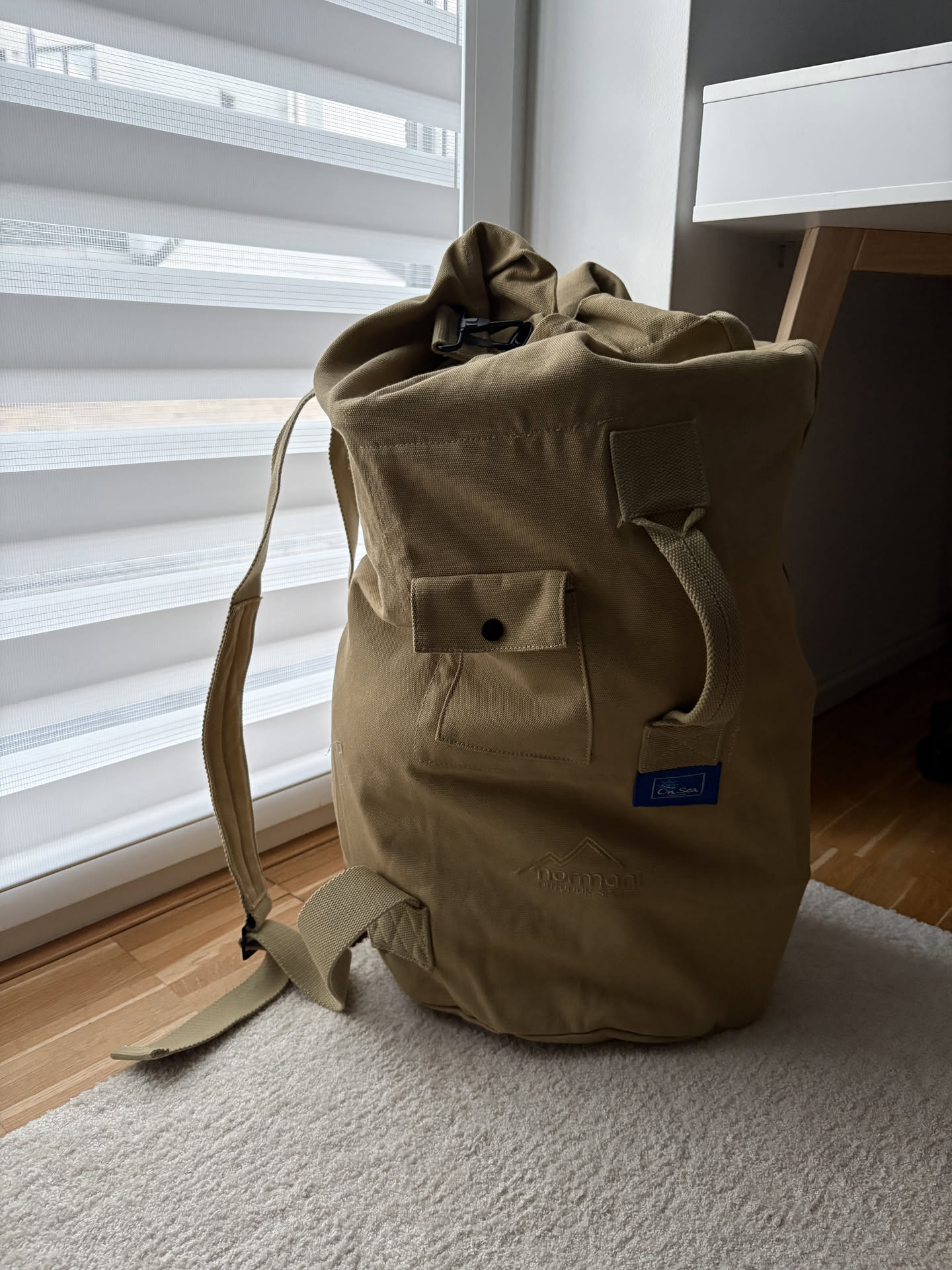 A khaki military canvas duffle bag standing on a carpet by a window with a roller blind. The bag has external pockets, carrying straps, and a top buckle closure.