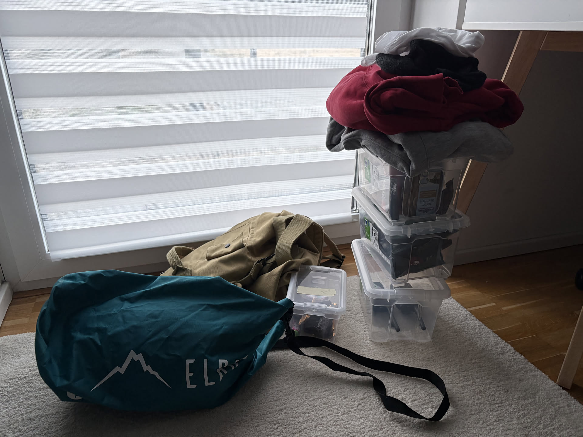 An evacuation gear set on a carpet by a window: a green bag with the Elbrus logo, a khaki military backpack, a small plastic container, and a stack of transparent clothing containers.